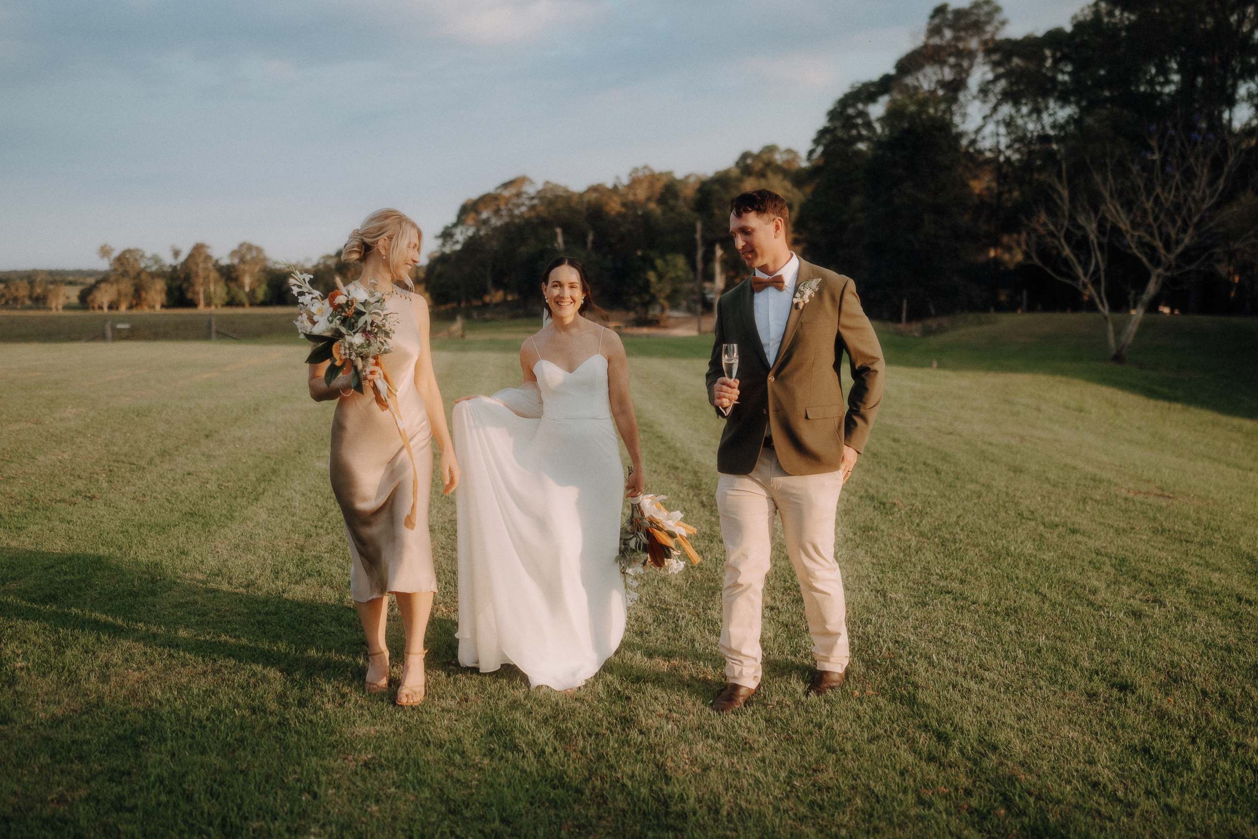 A bride in a white dress walks on grass with a bridesmaid in a gold dress and a groomsman in a suit, holding flowers and a champagne glass. Trees are visible in the background.