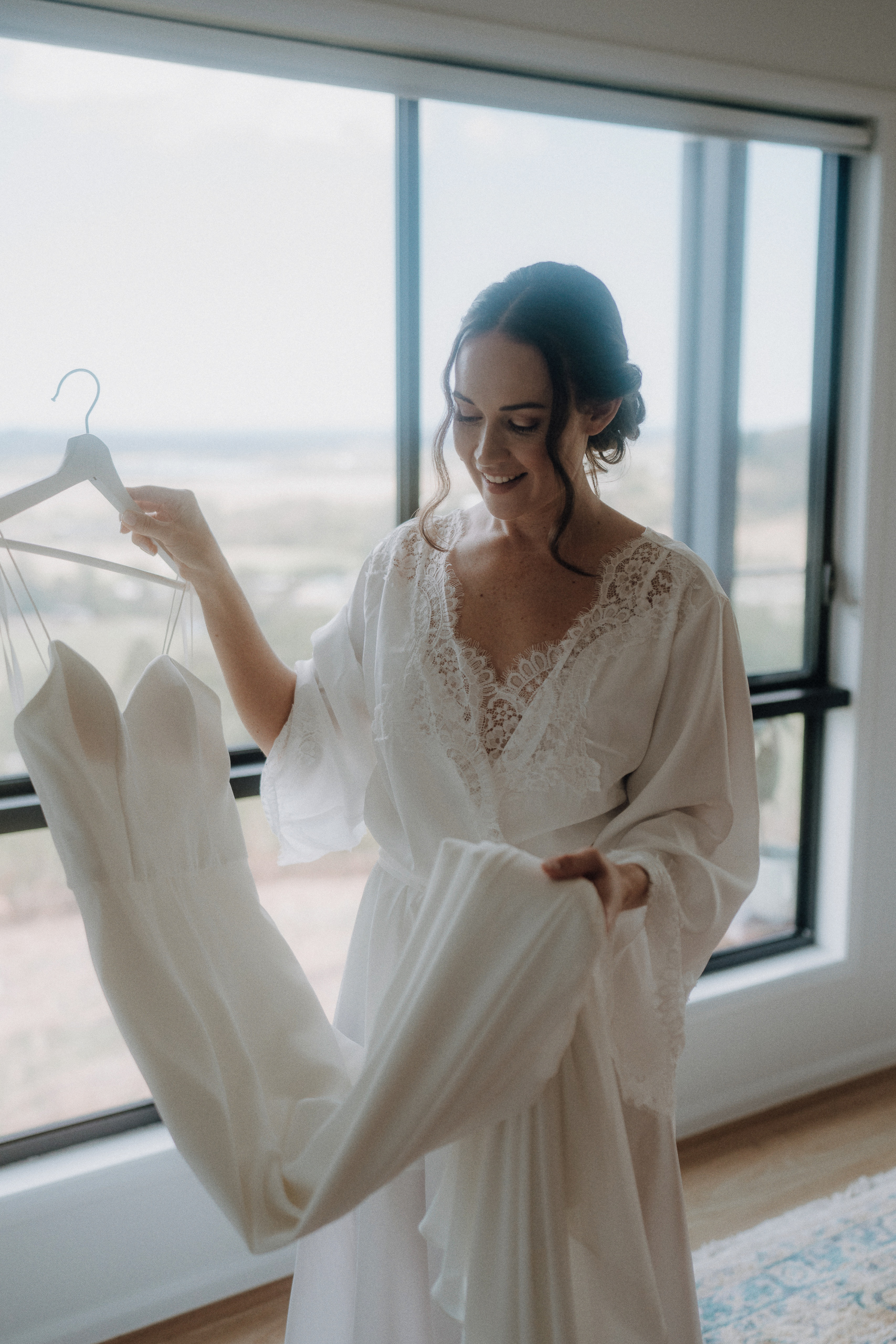 A woman in a white lace robe holds a wedding dress on a hanger, standing by a large window with an outdoor view.