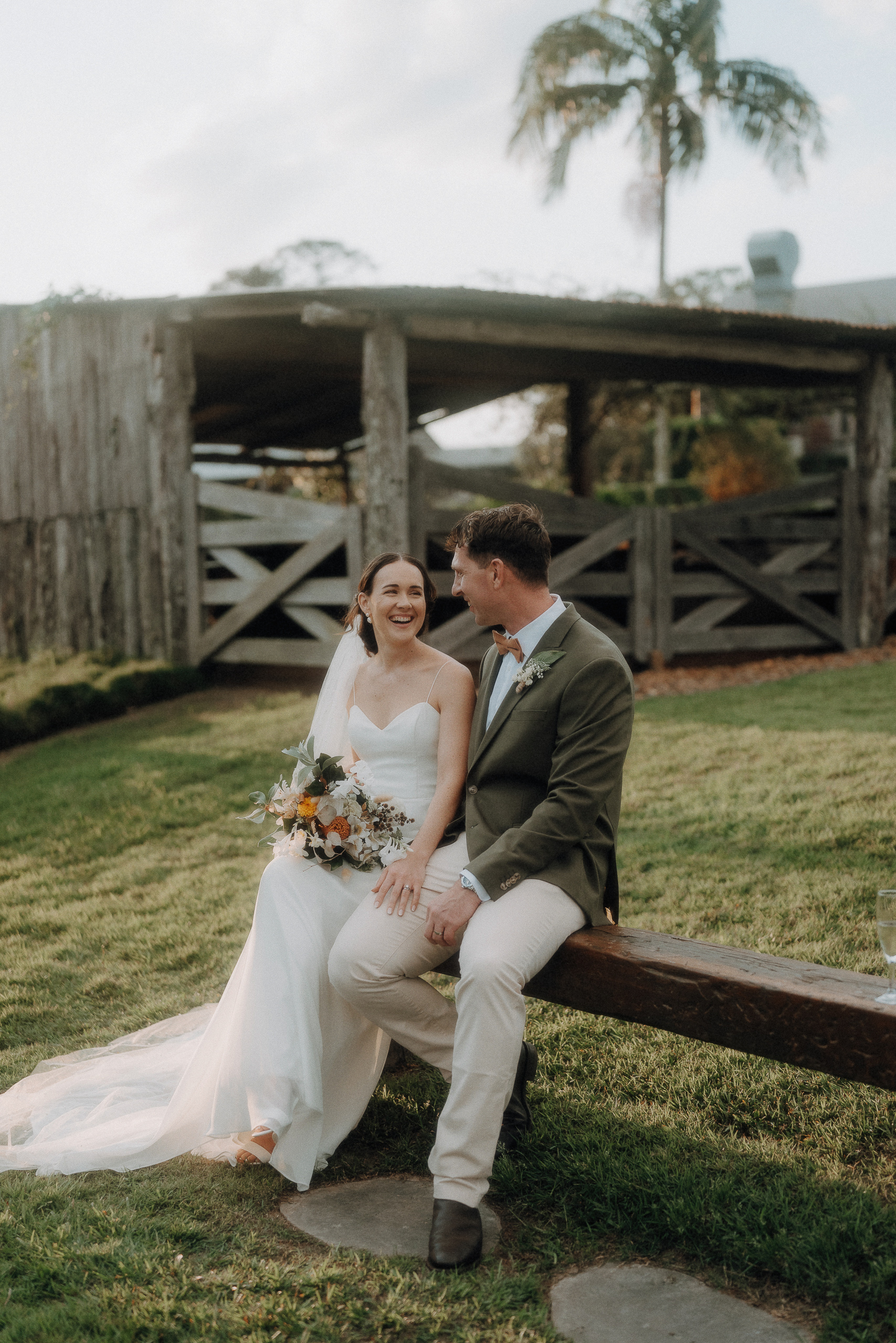 Bride and groom sit on a wooden beam outdoors, smiling at each other. The bride holds a bouquet, and a rustic wooden structure is visible in the background.