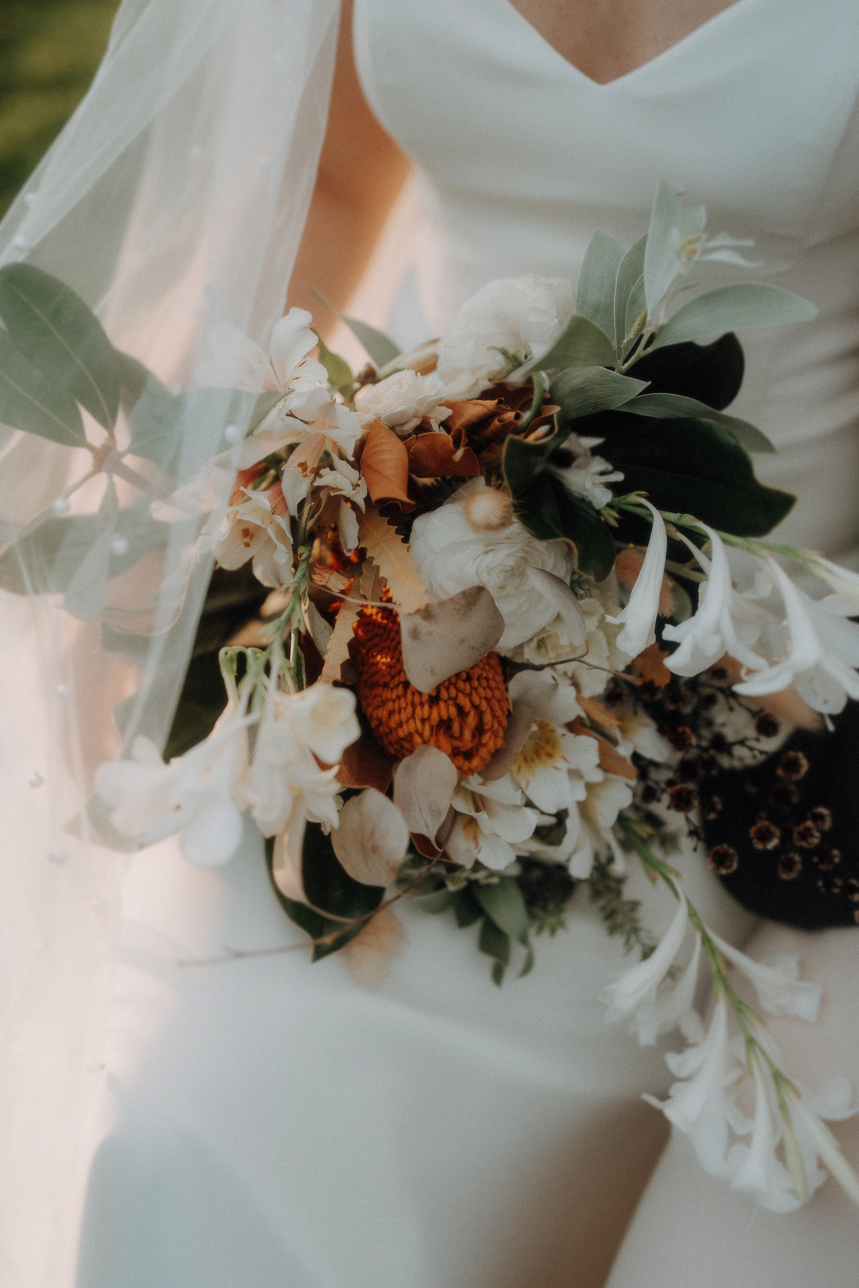 A bride in a white dress holds a bouquet of white flowers, green leaves, and some brown and orange floral accents.