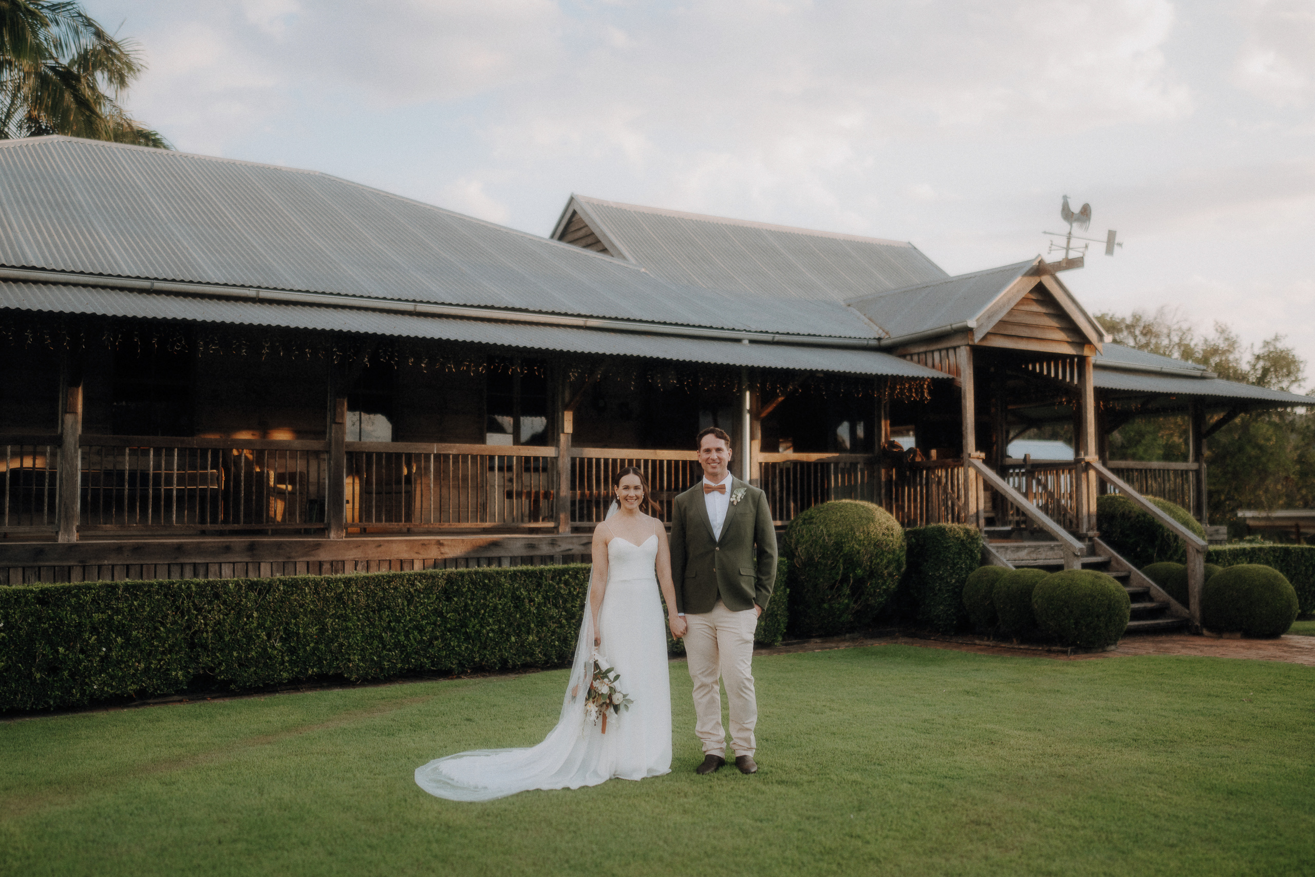 A bride in a white dress and a groom in a green jacket stand side by side on a lawn in front of a rustic wooden building.