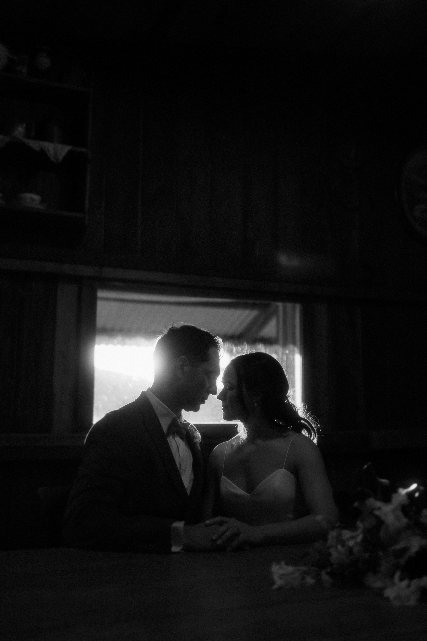 A couple sits closely together at a table in a dimly lit room, facing each other and holding hands, with light coming through a window behind them.