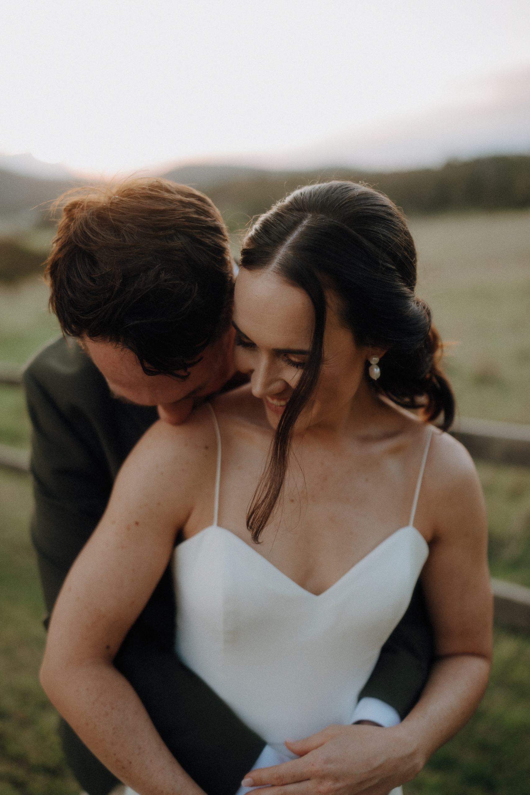 A couple embraces outdoors, with the man standing behind the woman in a white dress. They both smile and the background shows a blurred field and fence at sunset.