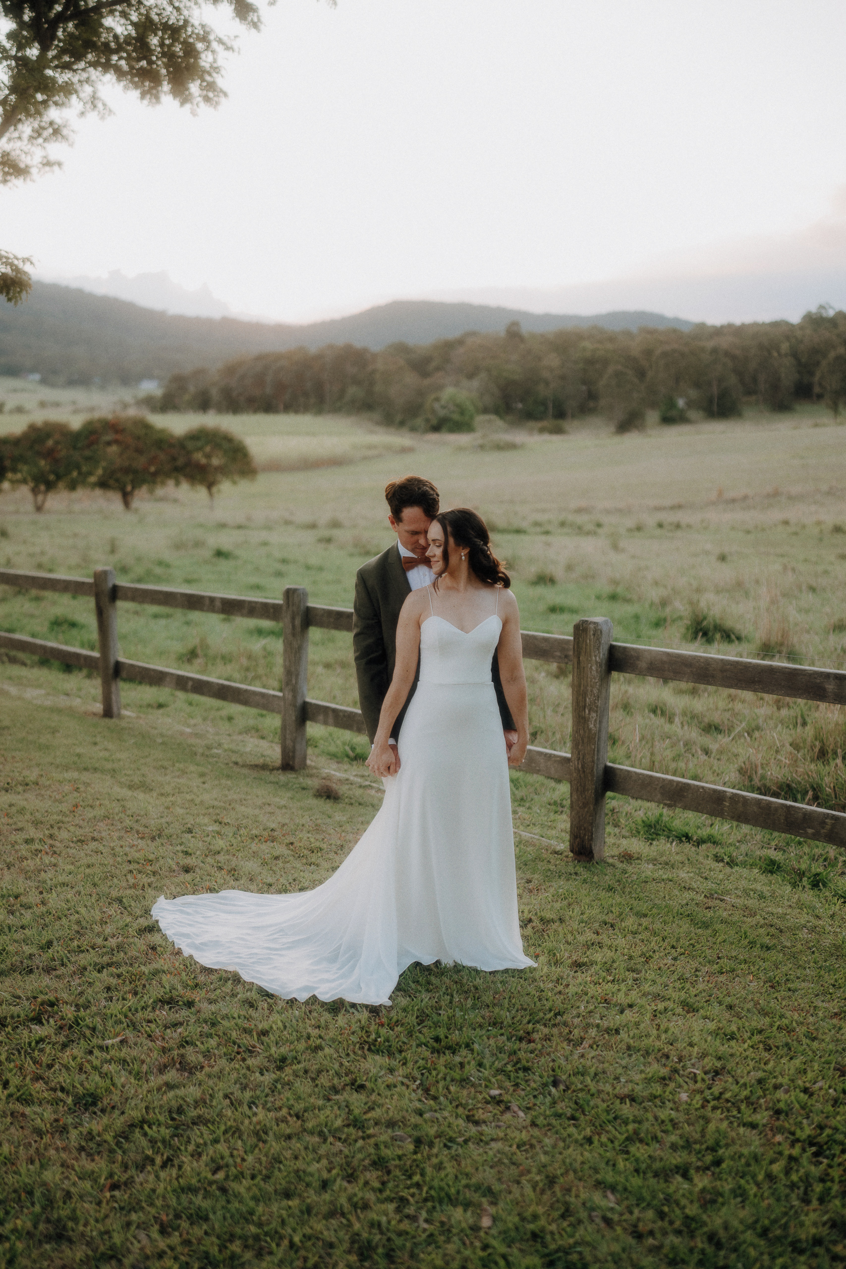 A couple in wedding attire stands on grass near a wooden fence in a scenic, rural landscape with hills and trees in the background.
