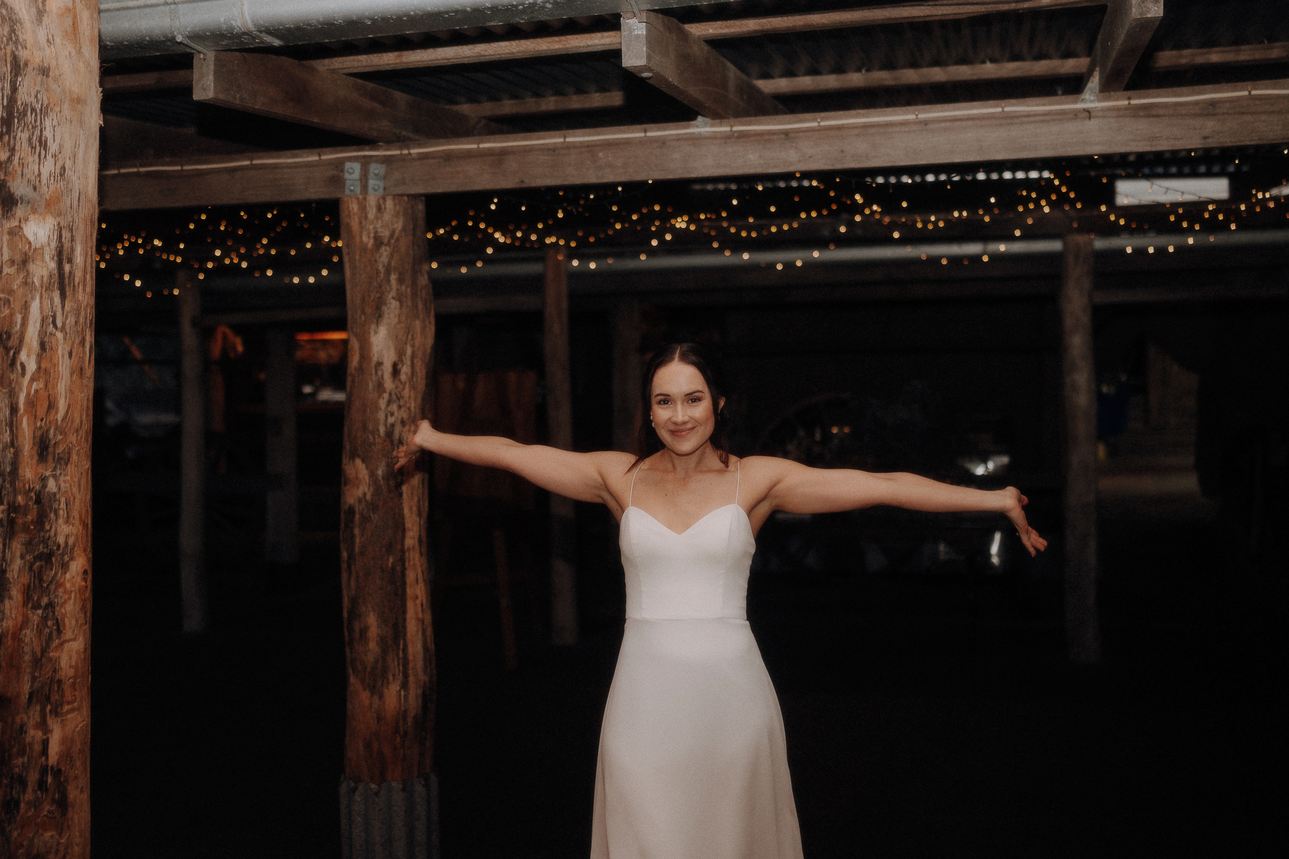 A woman in a white sleeveless dress stands indoors under string lights, extending both arms outward and smiling at the camera.