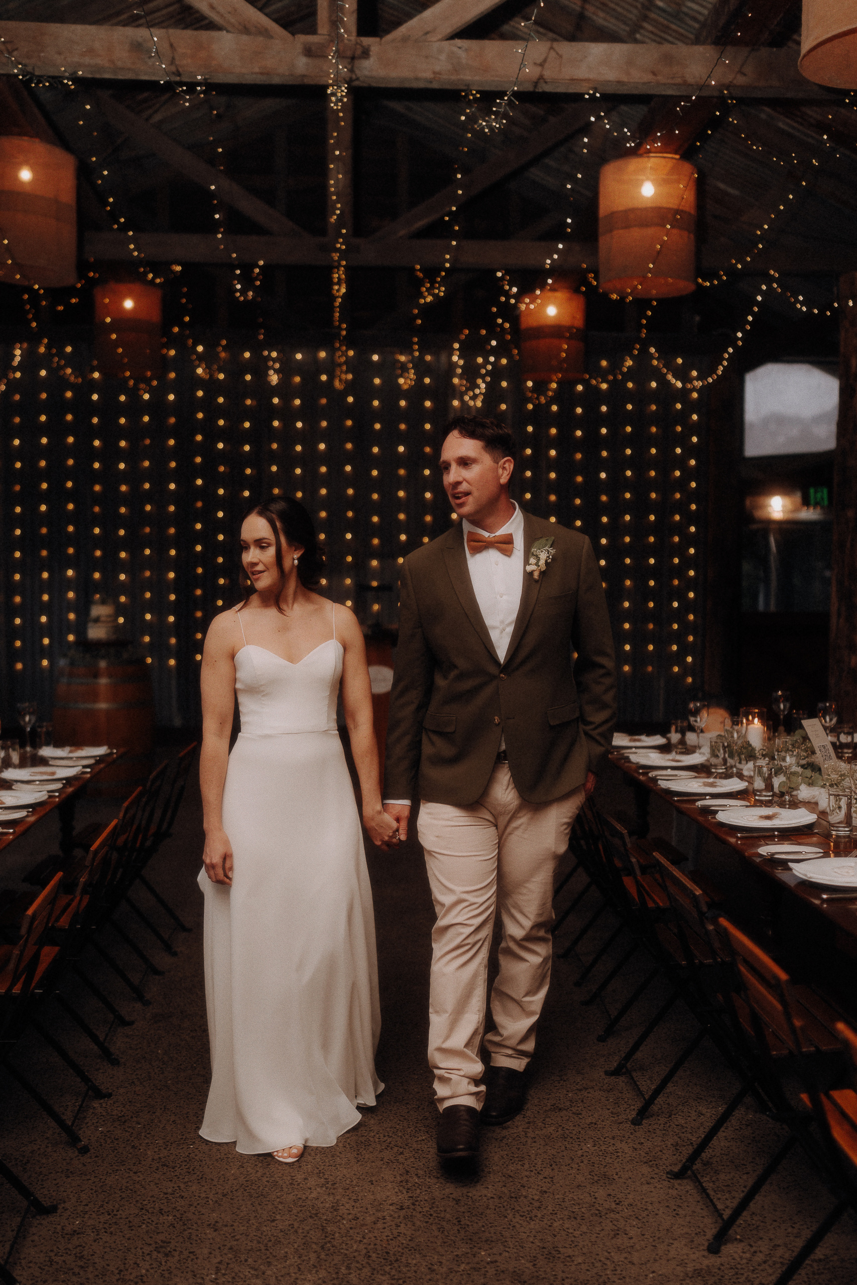 A couple in wedding attire walks hand in hand through a decorated venue with string lights and wooden beams overhead.
