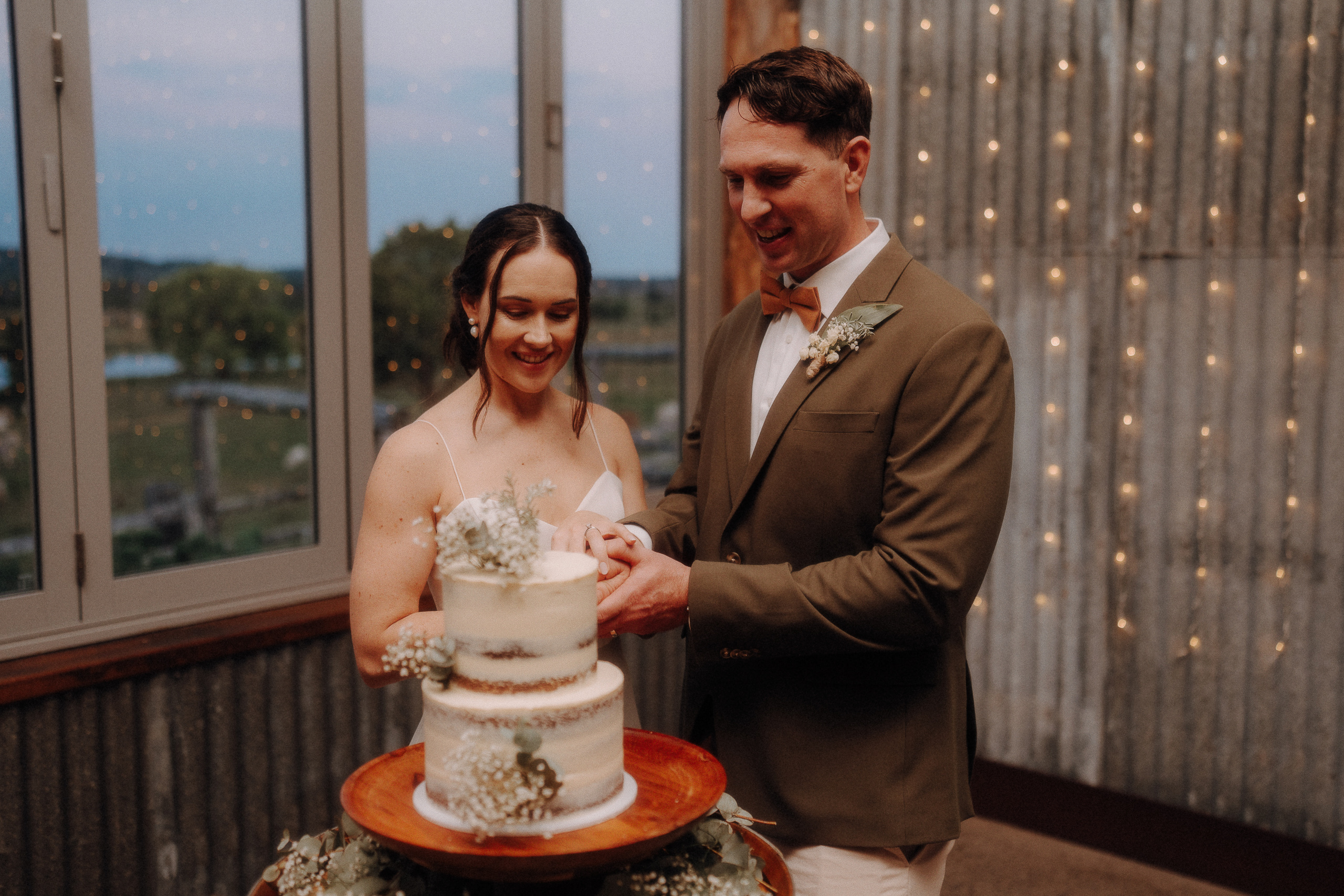 A bride and groom stand together, cutting a two-tiered white wedding cake decorated with flowers inside a rustic venue with string lights and large windows.