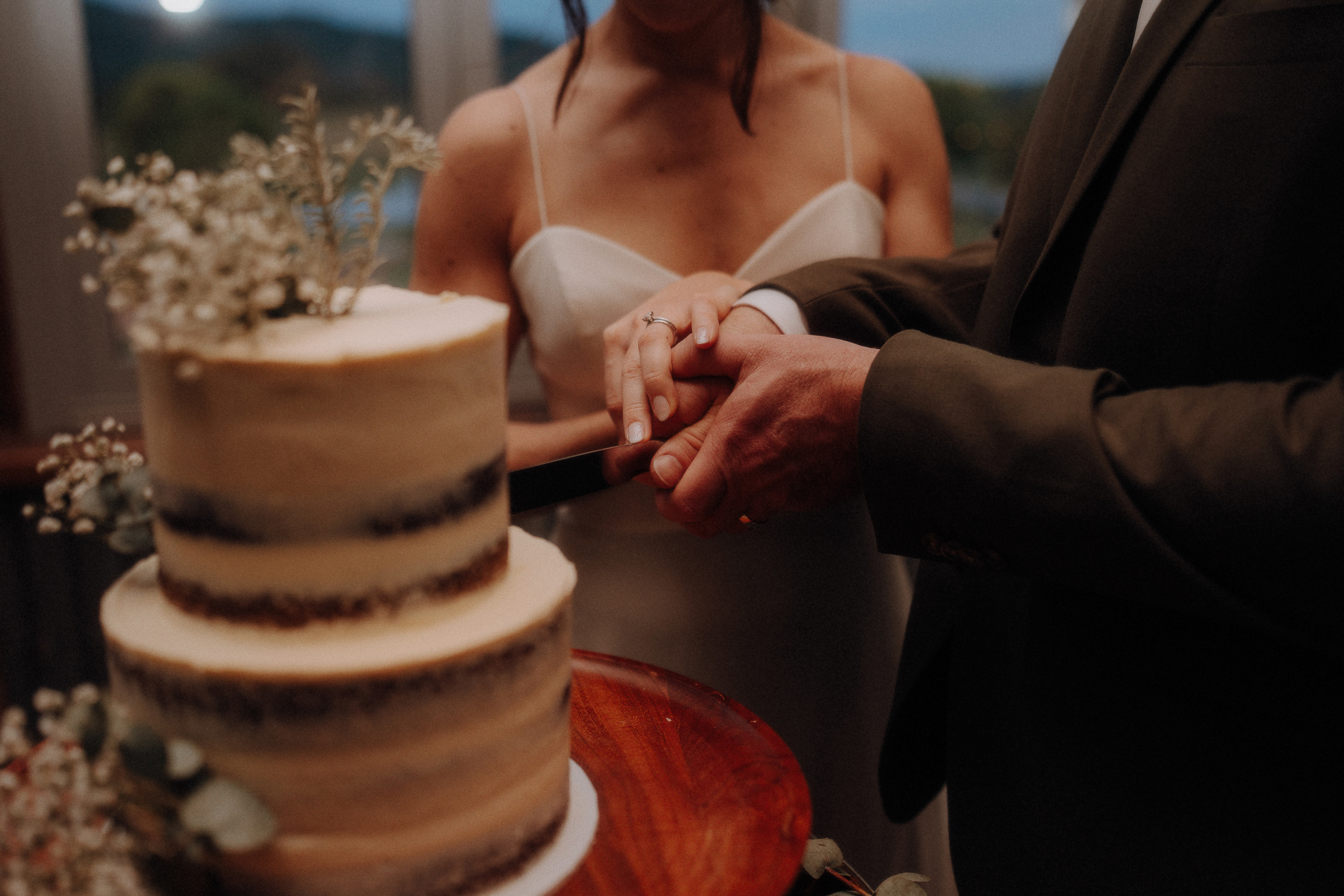 A couple, dressed formally, stands together as they cut a two-tiered rustic cake adorned with flowers.