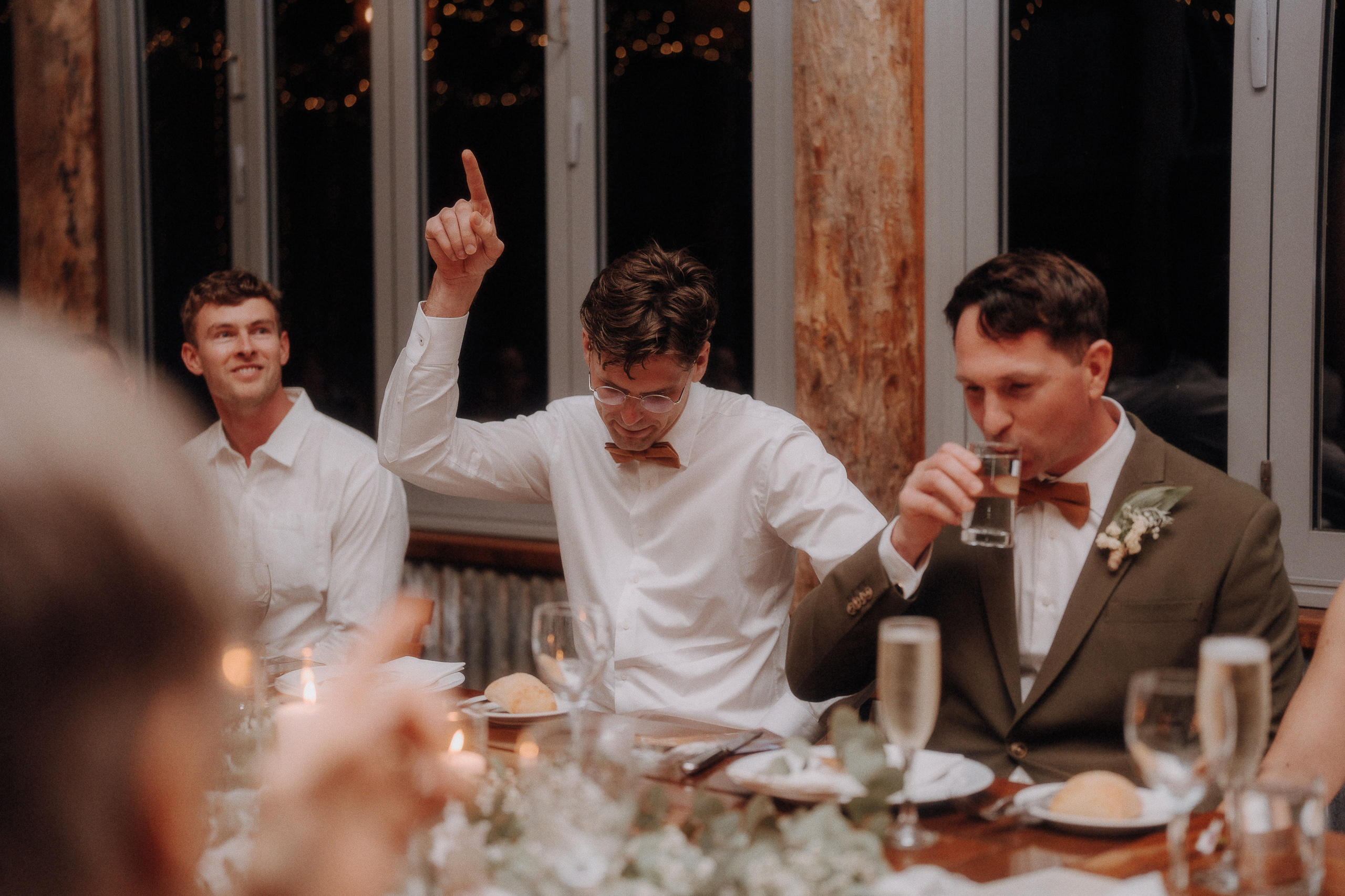 Three men sit at a dinner table; the man in the center raises one finger, the man on the right drinks water, and the man on the left looks forward. The table has glasses, bread, and candles.