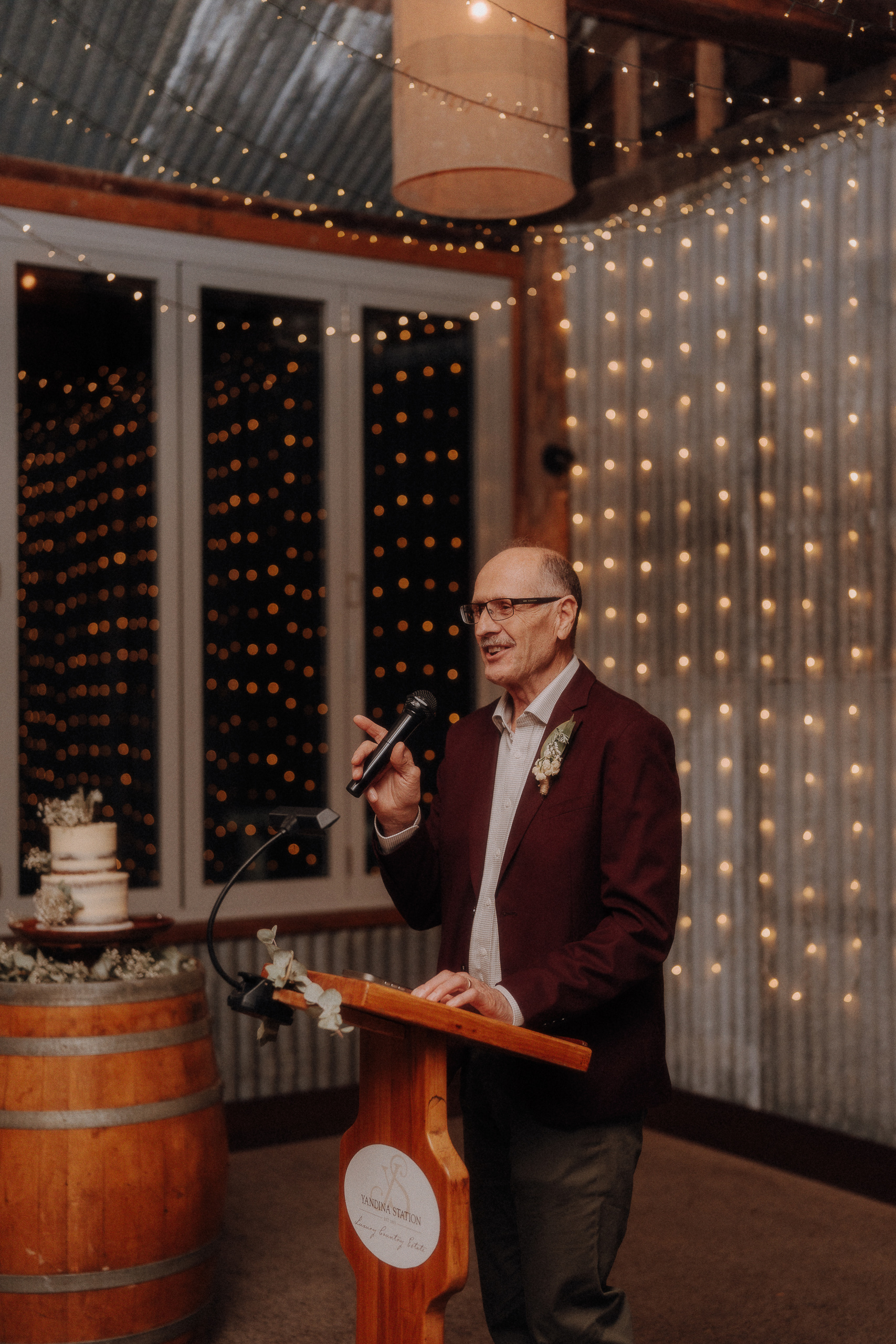 A man wearing glasses and a burgundy jacket speaks into a microphone at a podium, with string lights and a wedding cake on a barrel in the background.