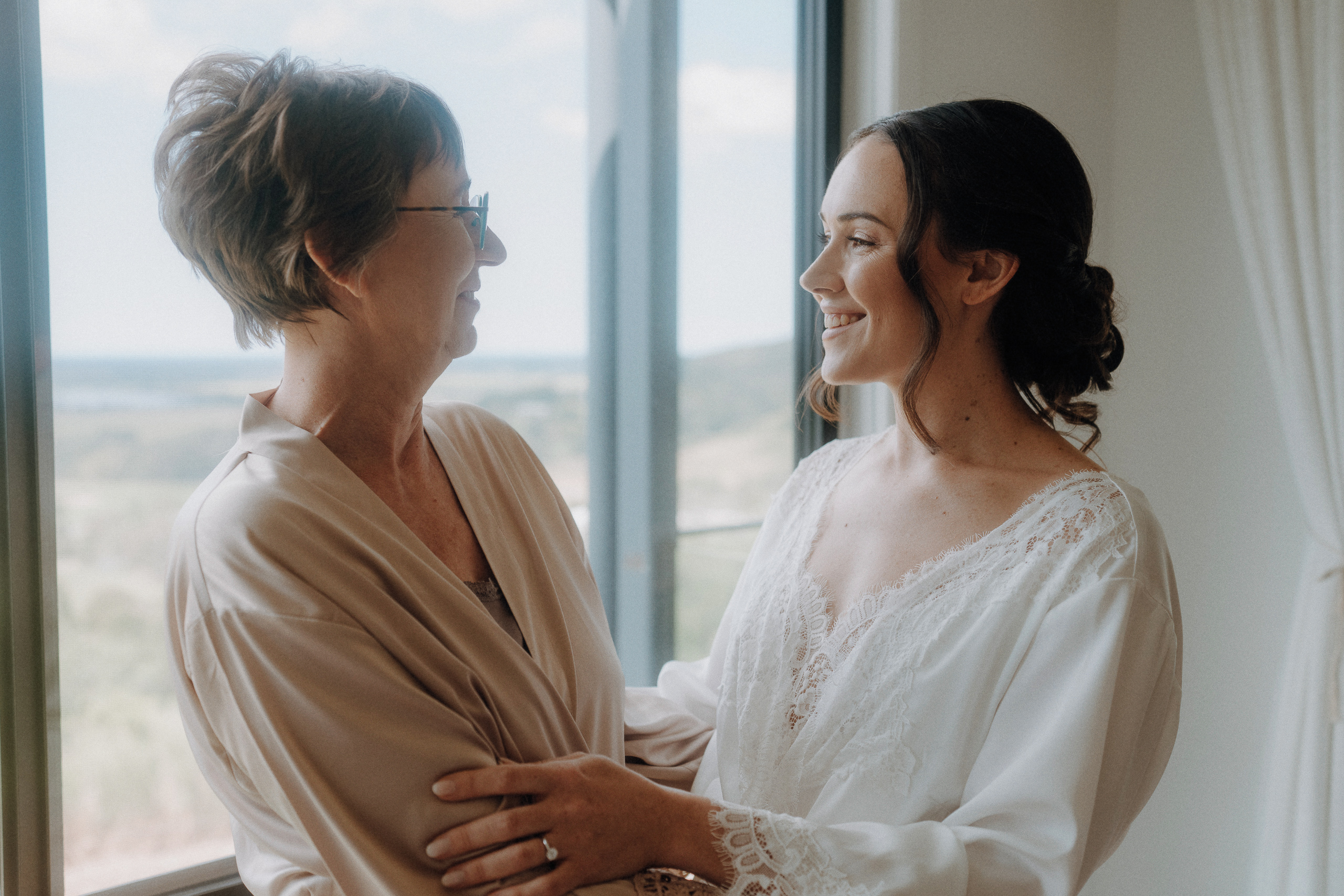 Two women in robes stand by a window, smiling at each other, with natural light illuminating their faces and a scenic view visible outside.