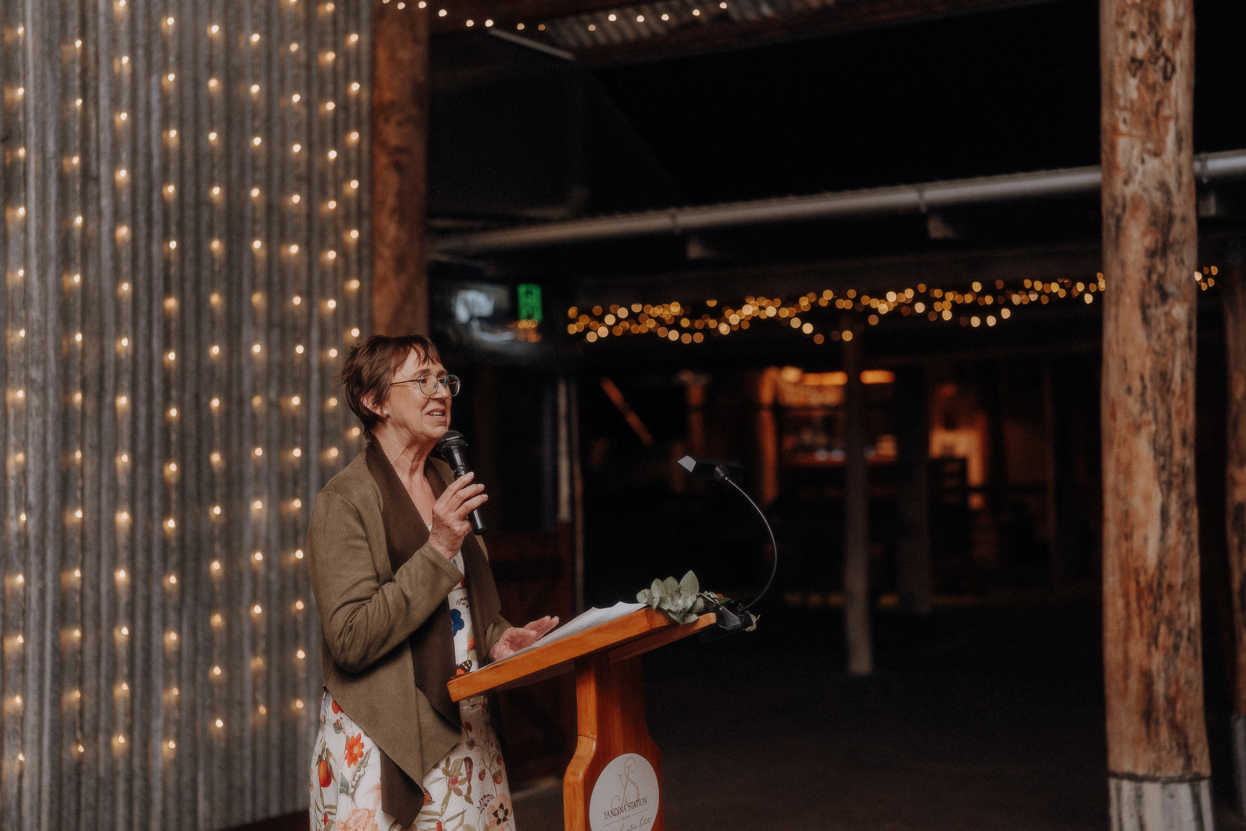 A woman stands at a wooden lectern, speaking into a microphone, with string lights hanging in the background of a dimly lit venue.