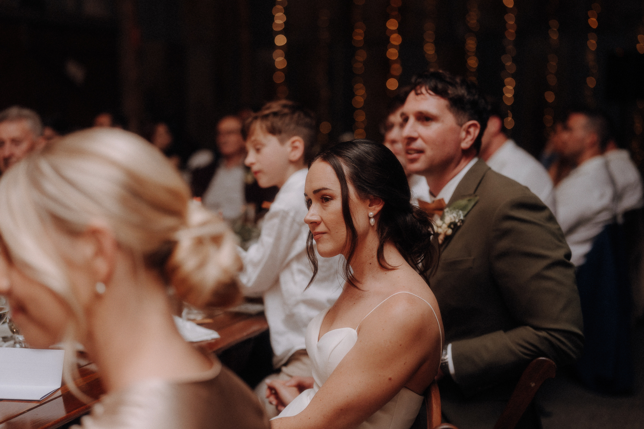 A bride and groom sit together at a table during a wedding reception, surrounded by guests, with warm string lights in the background.