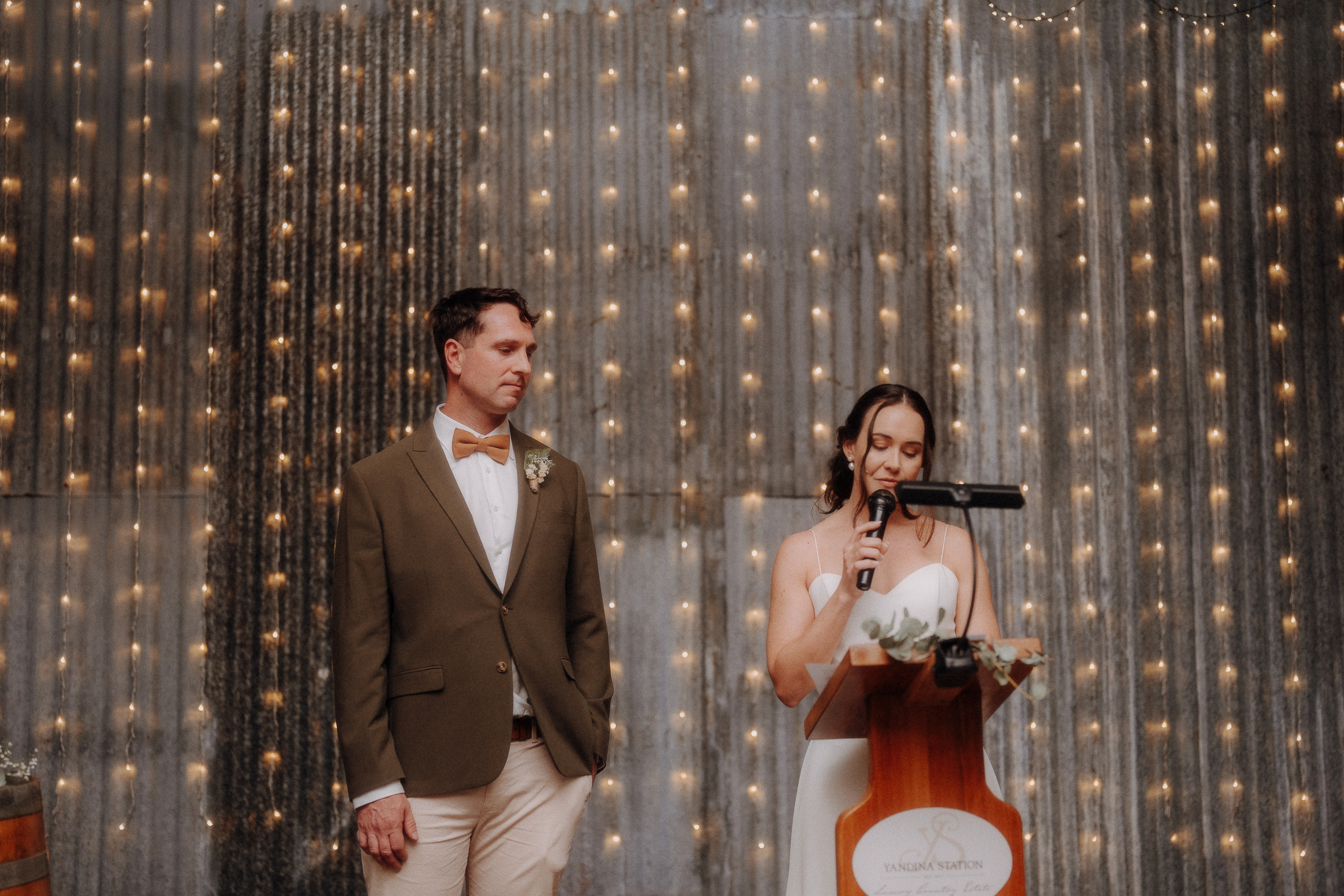 A woman in a white dress speaks at a podium with a microphone next to a man in a tan suit, in front of a curtain of string lights.