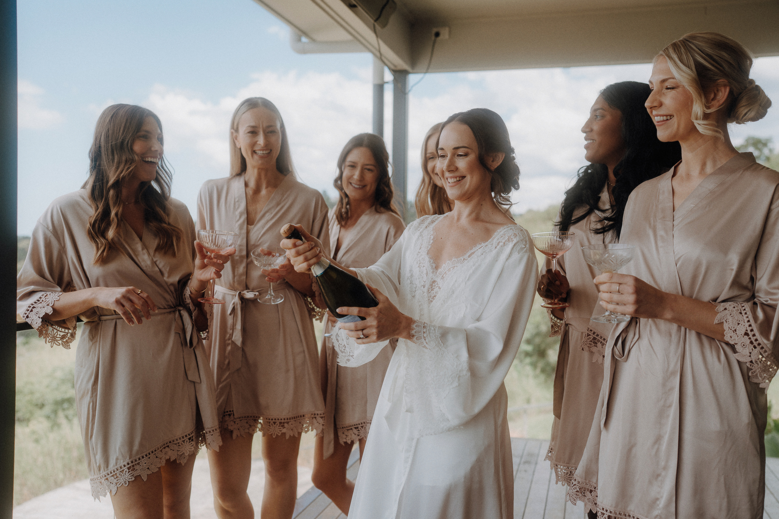 A bride in a white robe opens a bottle of champagne while five bridesmaids in matching robes stand around her holding glasses on a porch.
