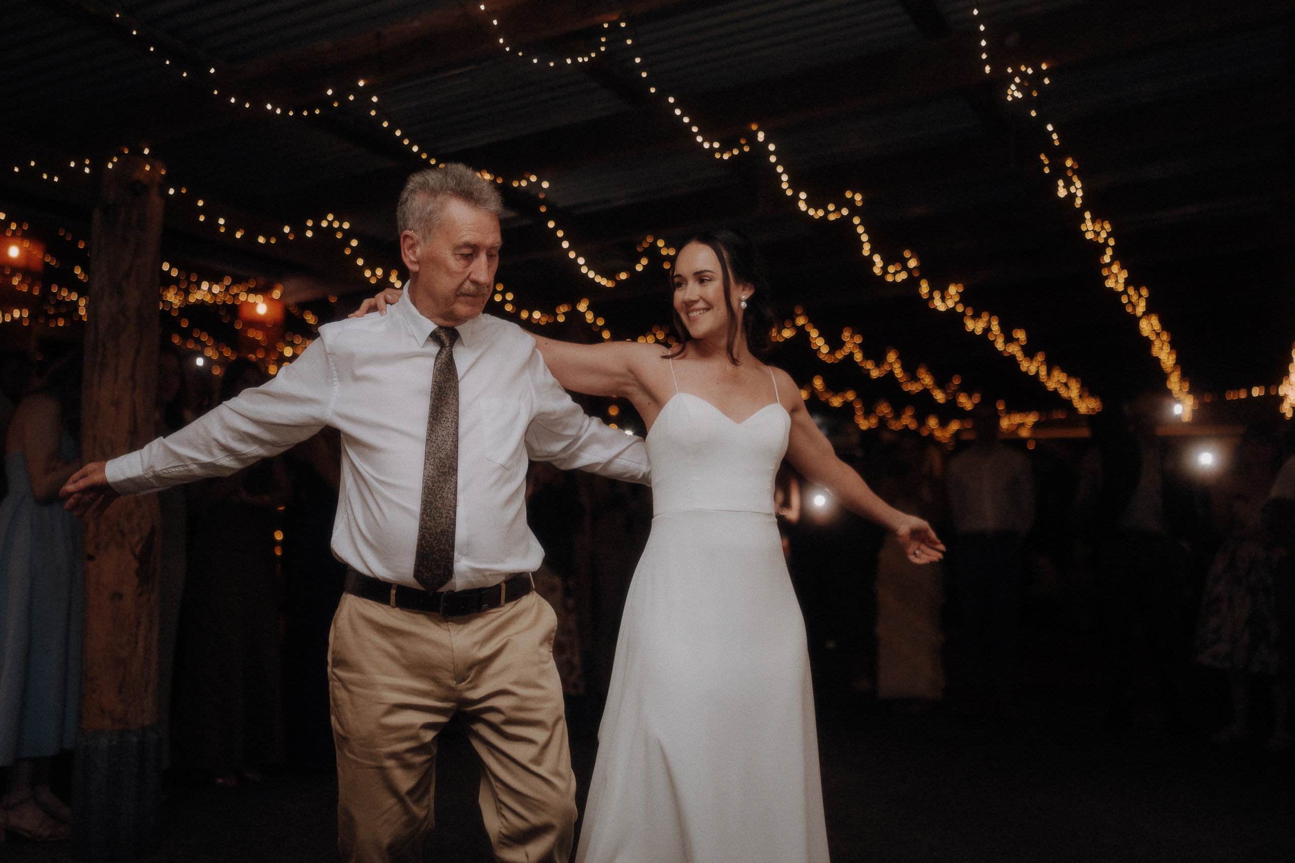 A man in a white shirt and tie dances with a woman in a white dress under string lights at a dimly lit event.