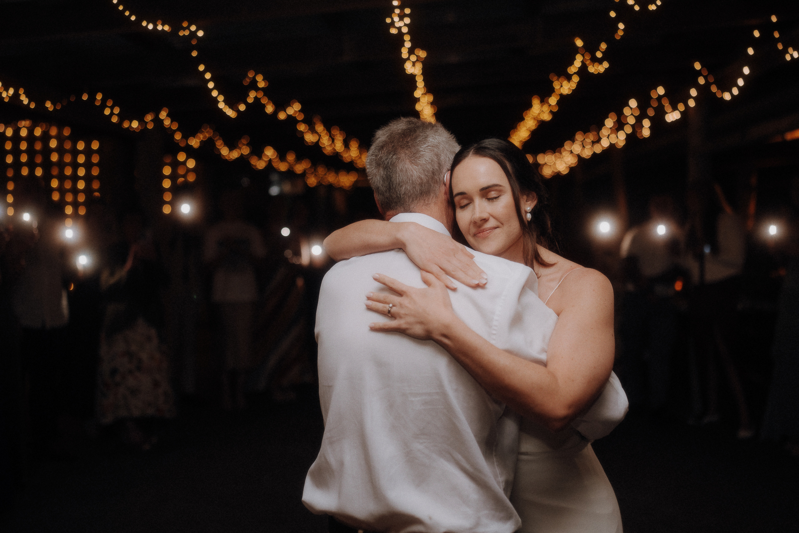 A bride with closed eyes hugs an older man in a white shirt while dancing under string lights at a nighttime event, surrounded by people holding up phone lights.