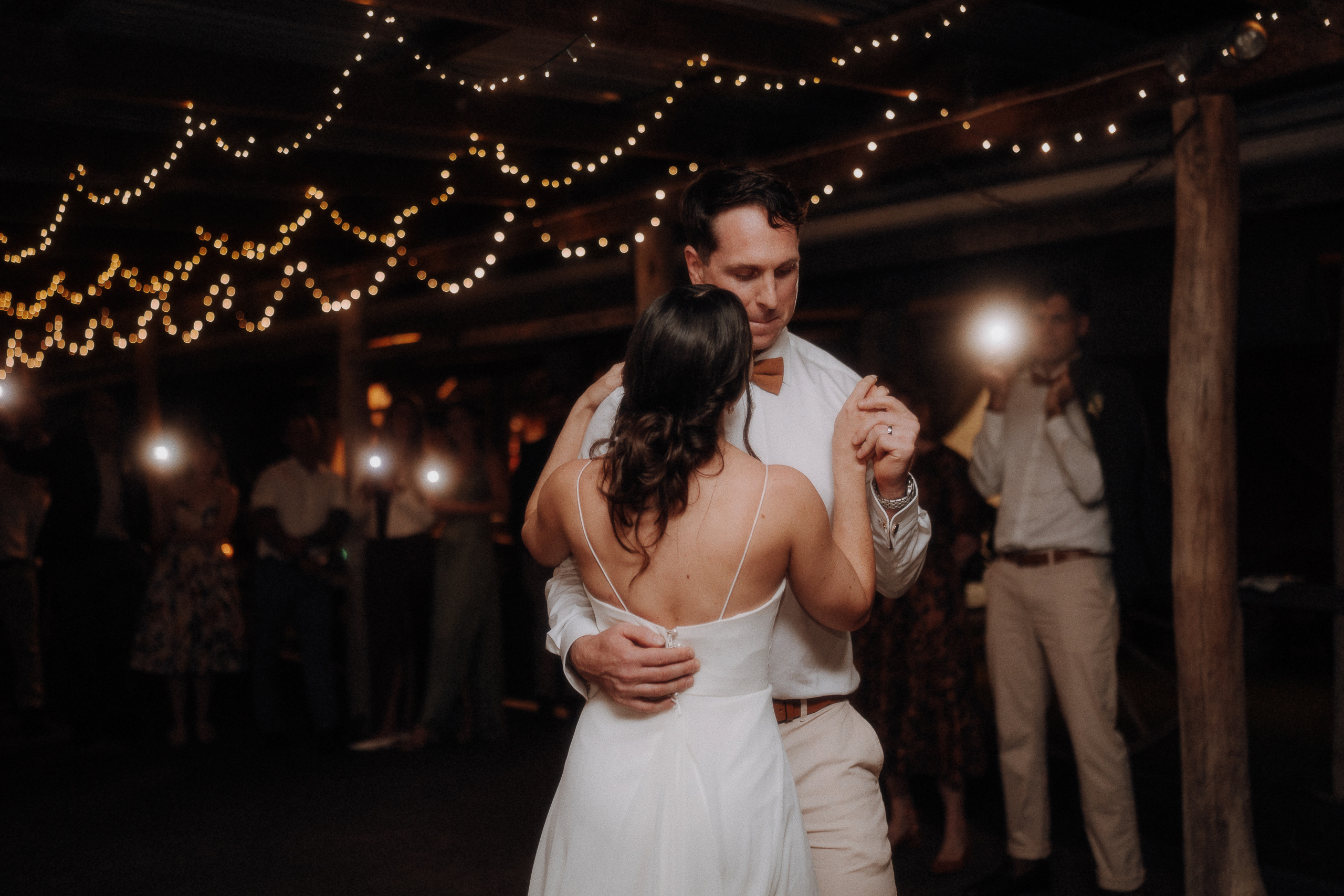 A couple in wedding attire shares a first dance under string lights, with guests in the background holding up phones to capture the moment.