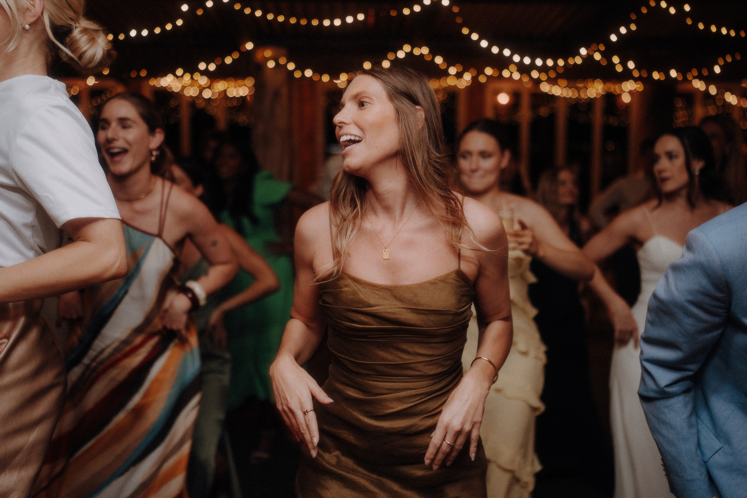 A group of people dance at an indoor event under string lights. A woman in the foreground wears a brown dress and smiles, surrounded by others in formal attire.