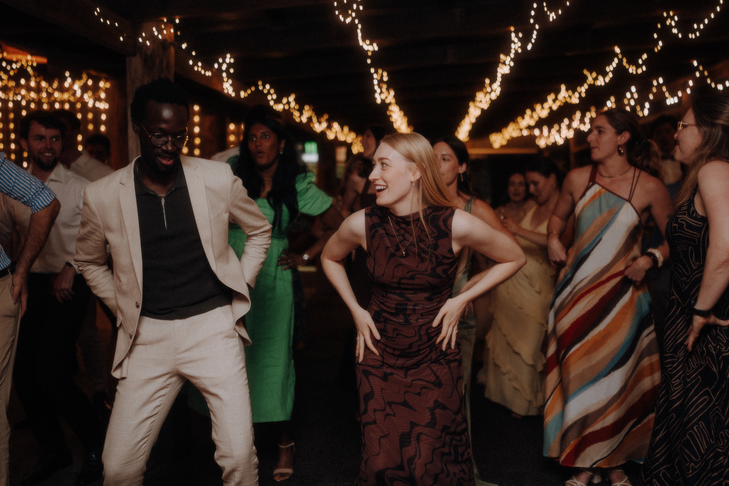 A group of people dance together under string lights at an indoor event, with two people in the front smiling and leading the group.