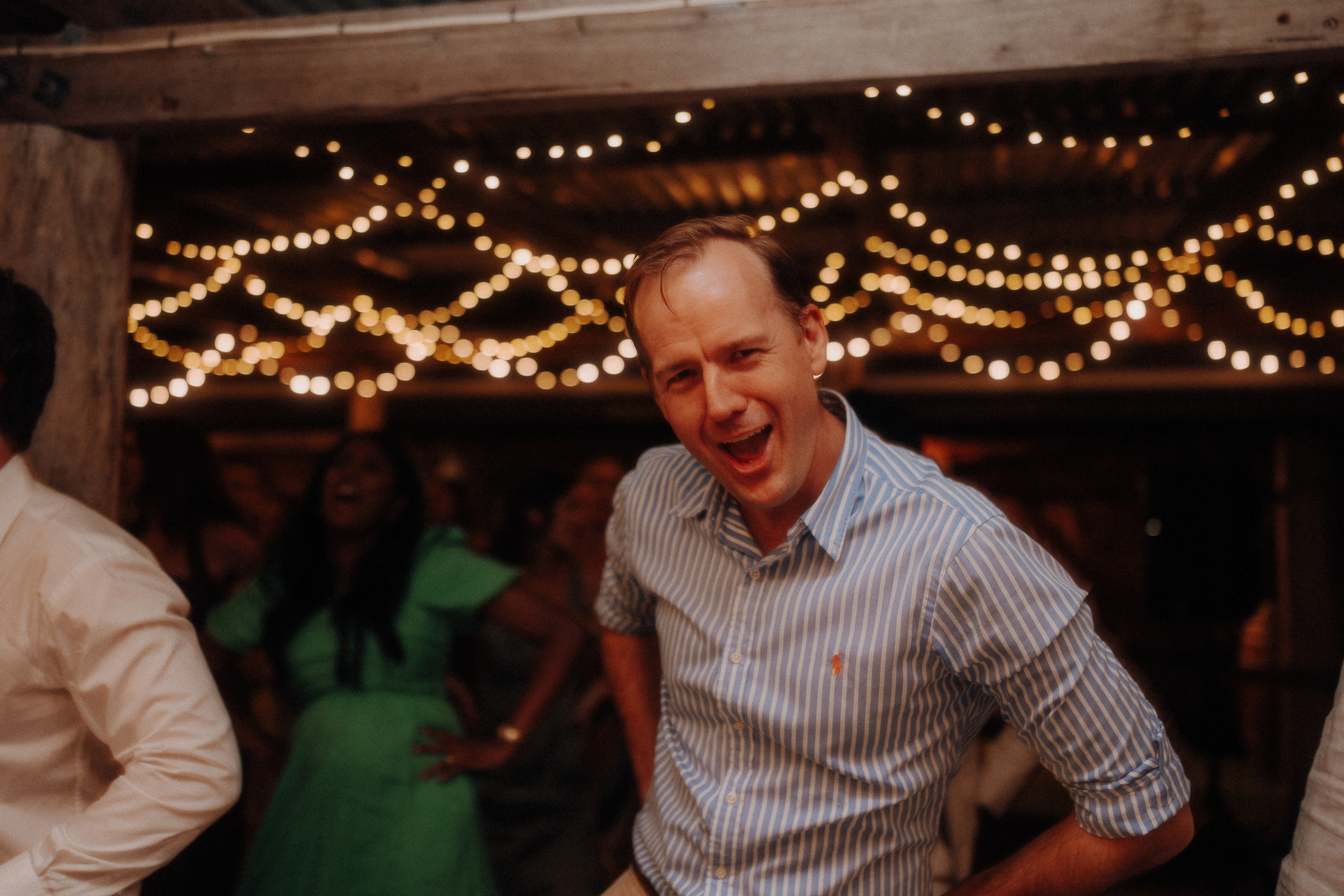 A man in a blue and white striped shirt poses energetically at an indoor event with string lights and people in the background.