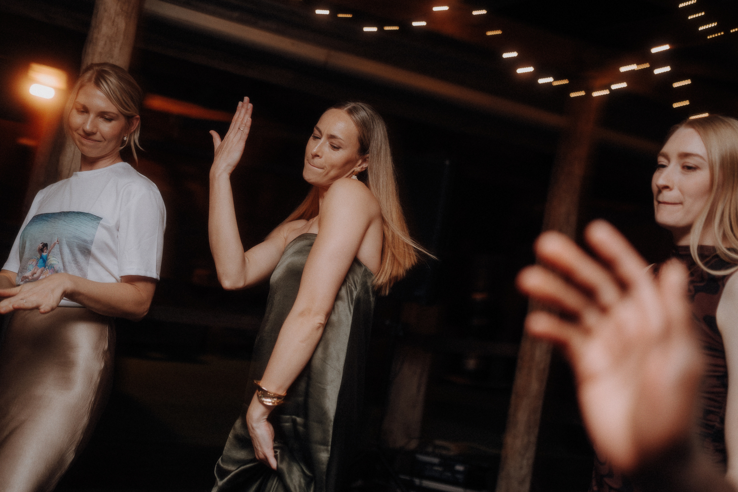 Three women are dancing indoors under string lights, with one woman raising her hand while the others look on and smile.