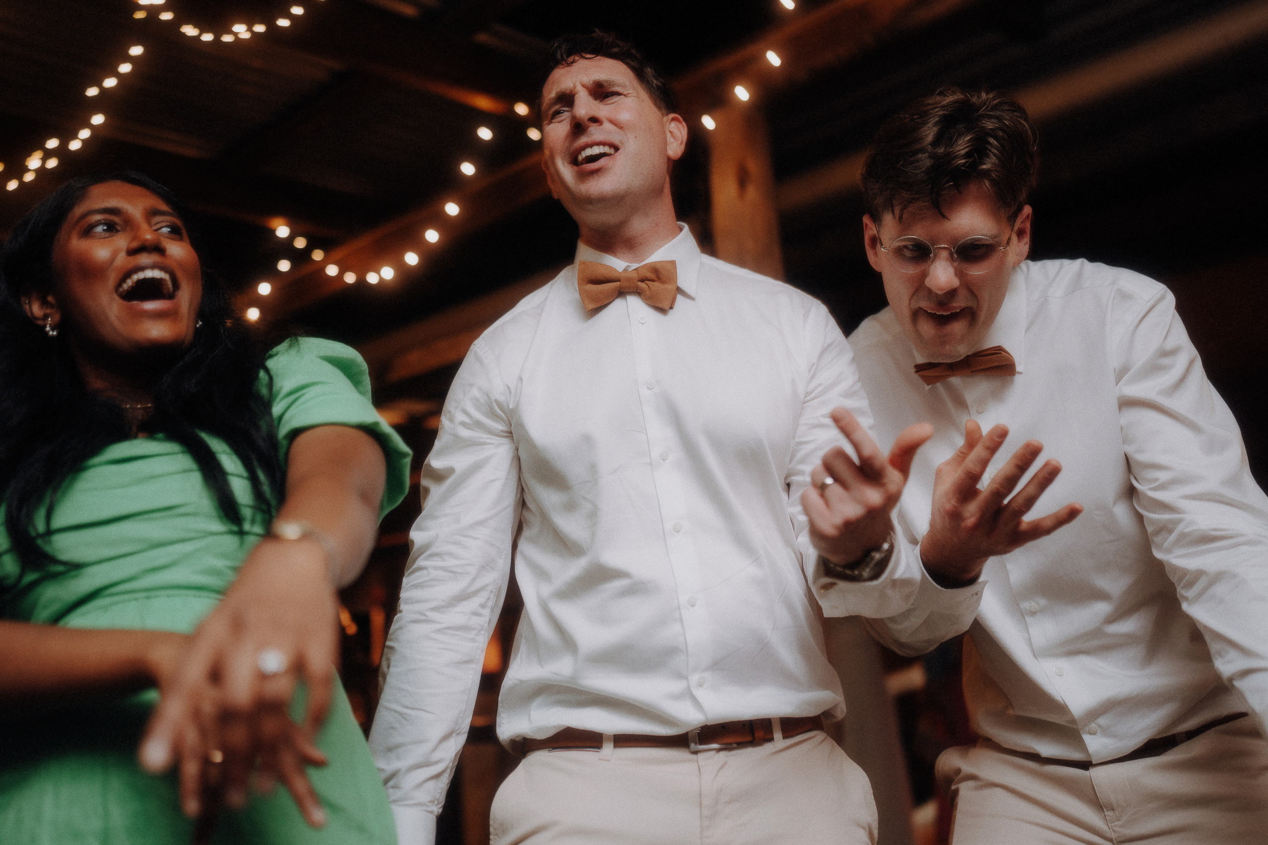 Three people dressed in formal attire enthusiastically sing and gesture at an indoor event with string lights in the background.