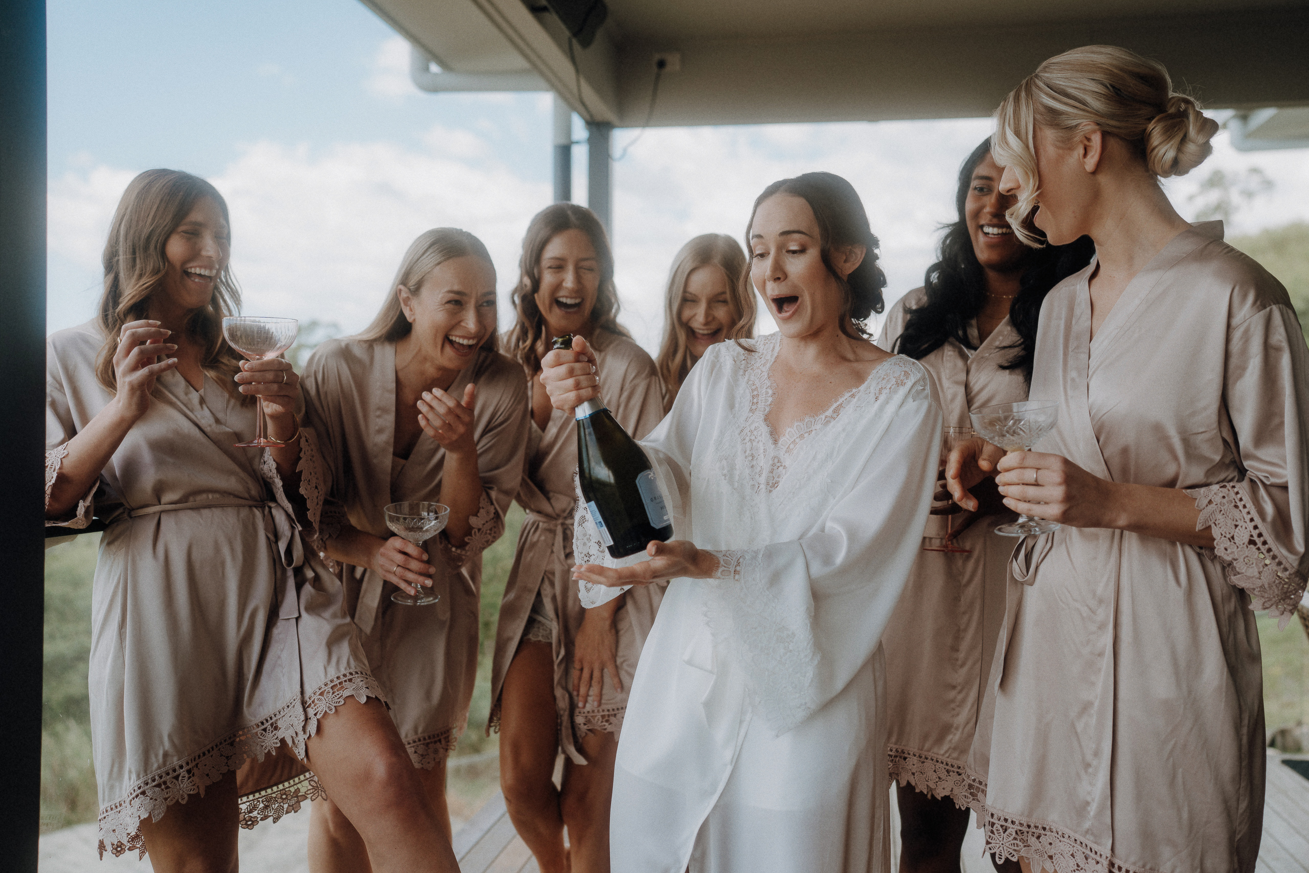 A bride in a white robe opens a bottle of champagne surrounded by six bridesmaids in matching beige robes, all smiling and holding glasses.