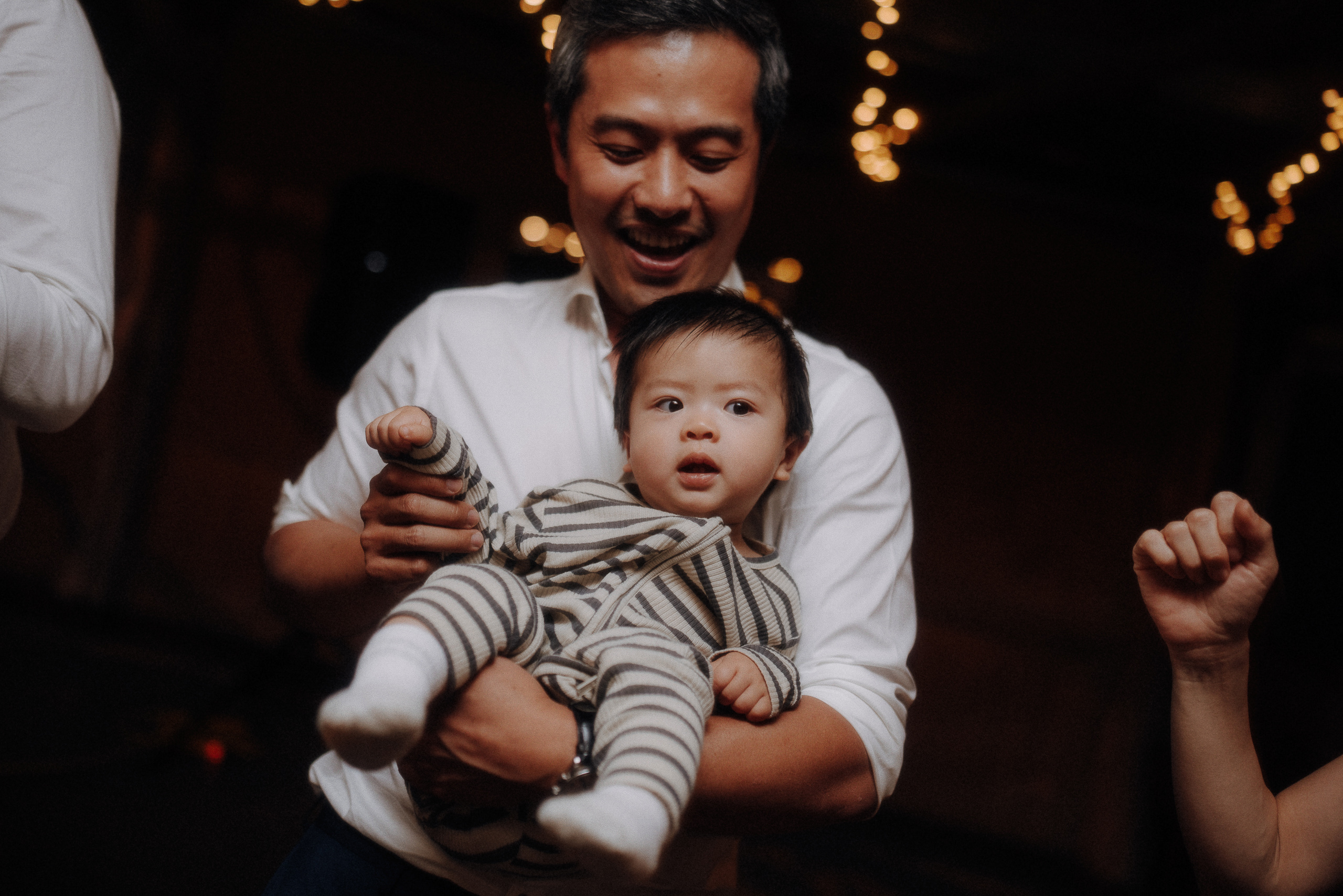 An adult holding a baby wearing striped pajamas, both appear to be enjoying themselves under string lights.