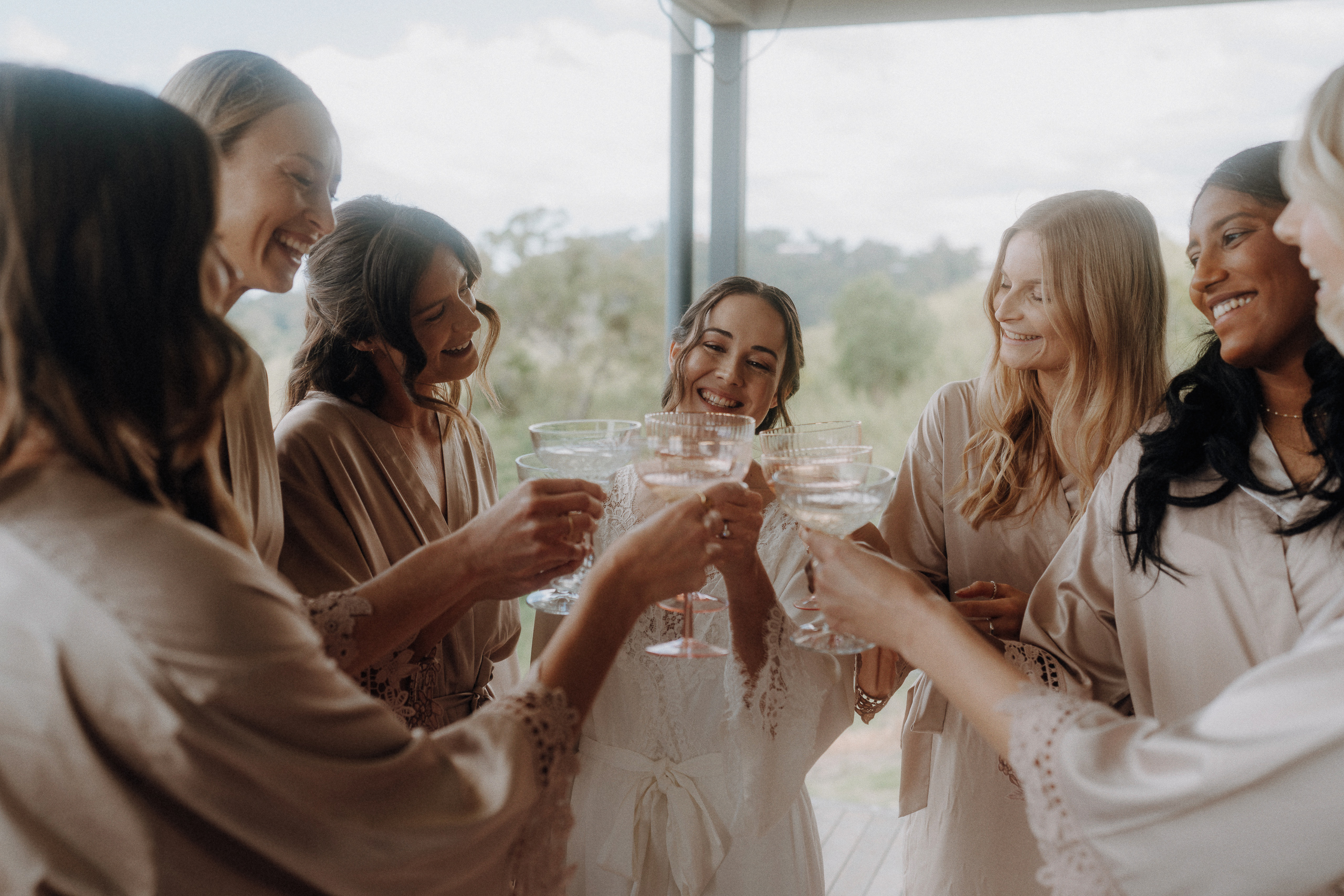 Seven women in matching robes smile and toast with glasses, standing together in a bright, outdoor setting.