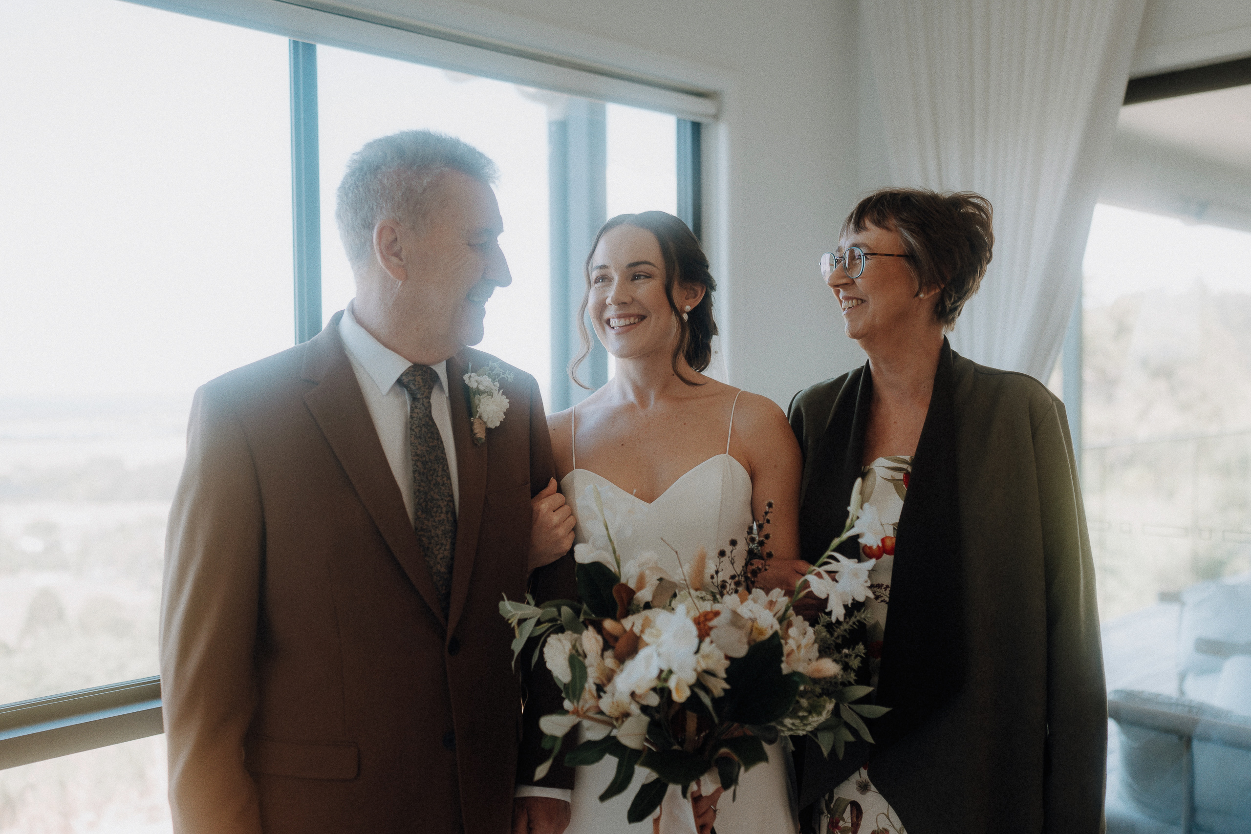 A bride holding a bouquet stands smiling between two adults in formal attire by a window in a bright room.