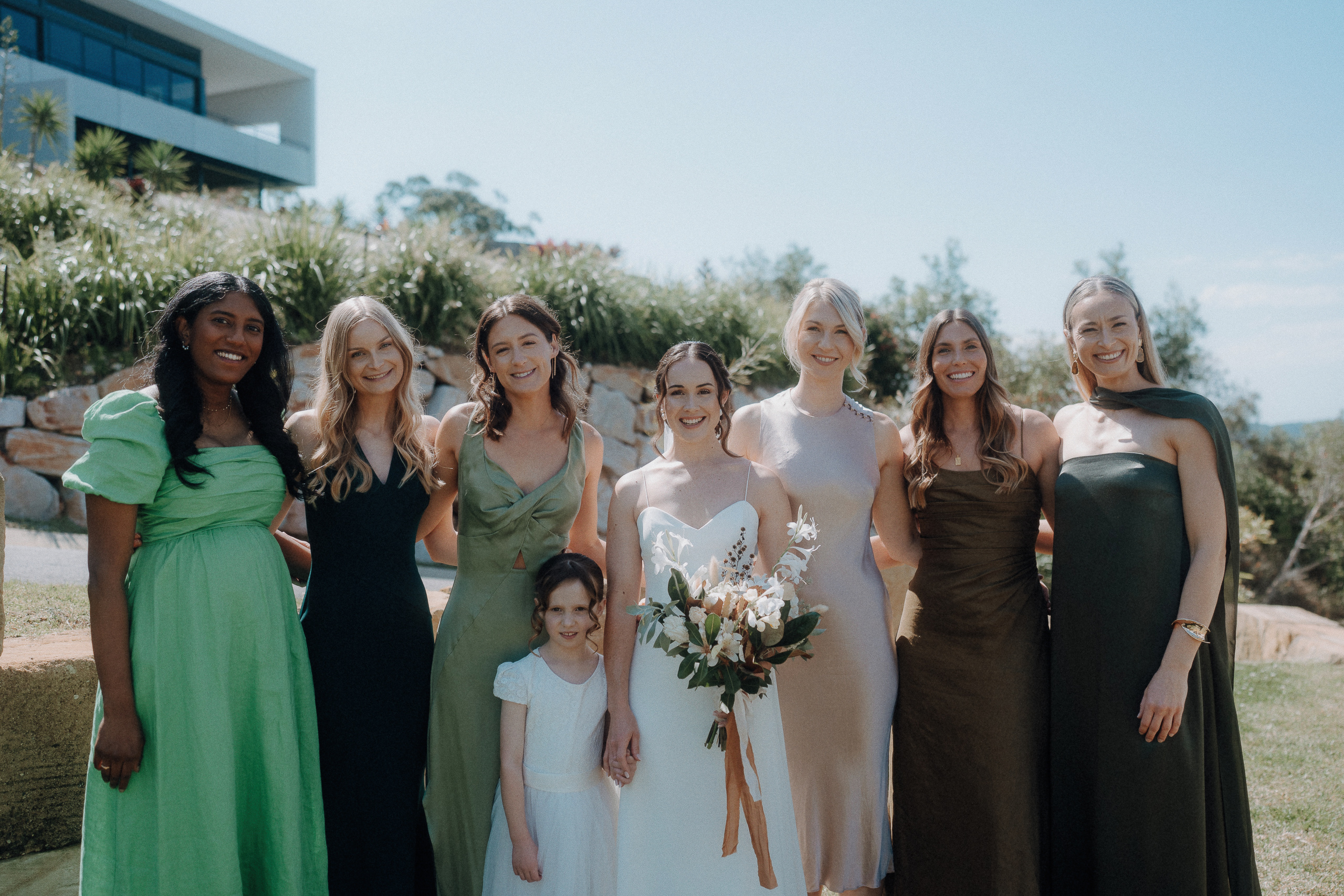 A bride holding a bouquet stands with six bridesmaids and a young flower girl outdoors on a sunny day, with greenery and a modern building in the background.