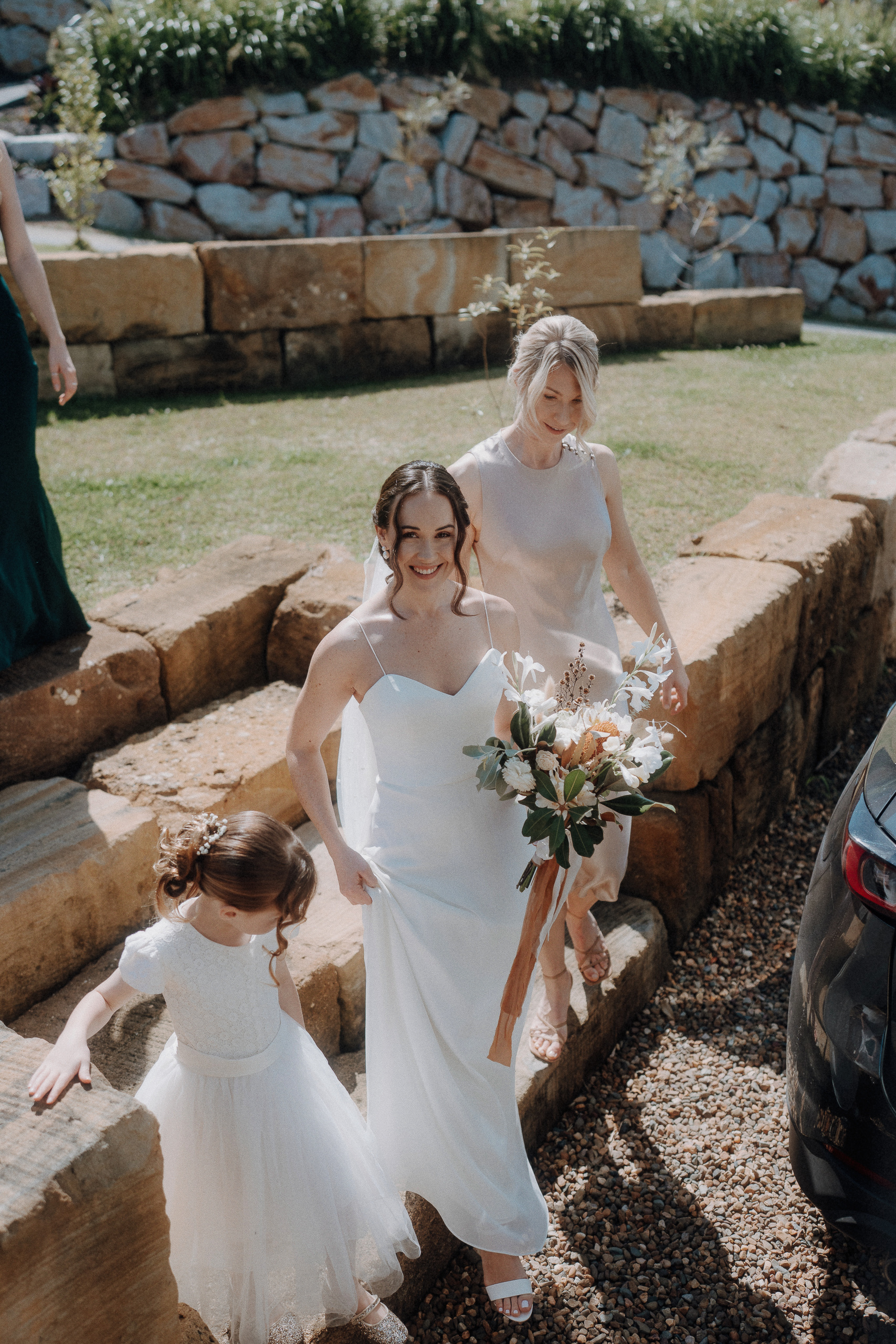 A bride in a white dress holds a bouquet and walks outside with a bridesmaid and a young flower girl on a stone path.